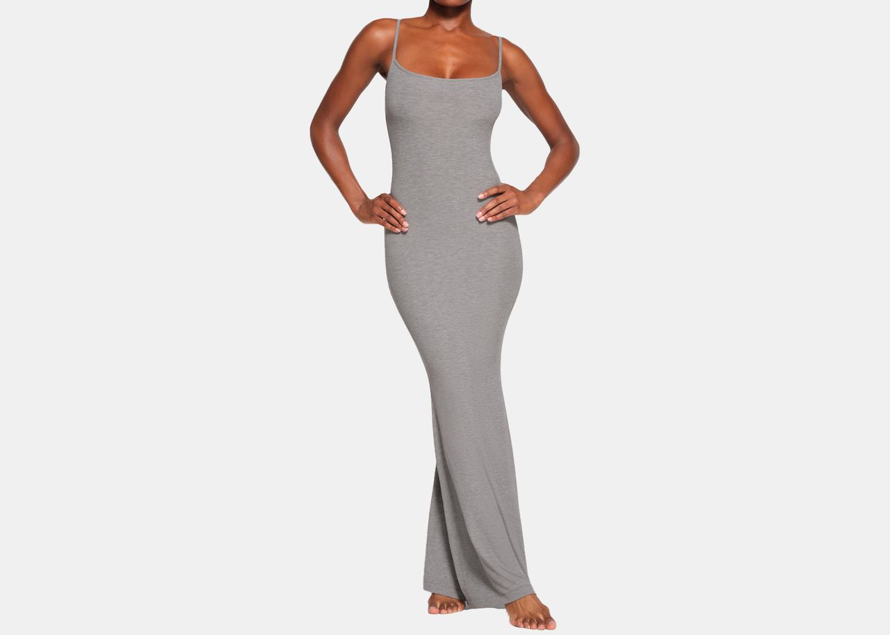 Female model in long, grey, slimming dress with hands on hips and one leg crossed over the other.