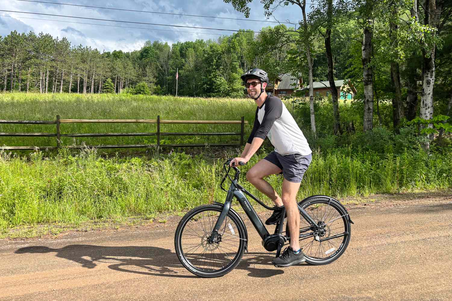 A man riding a Priority electric bike on a dirt road, with a wooden fence and trees in the background