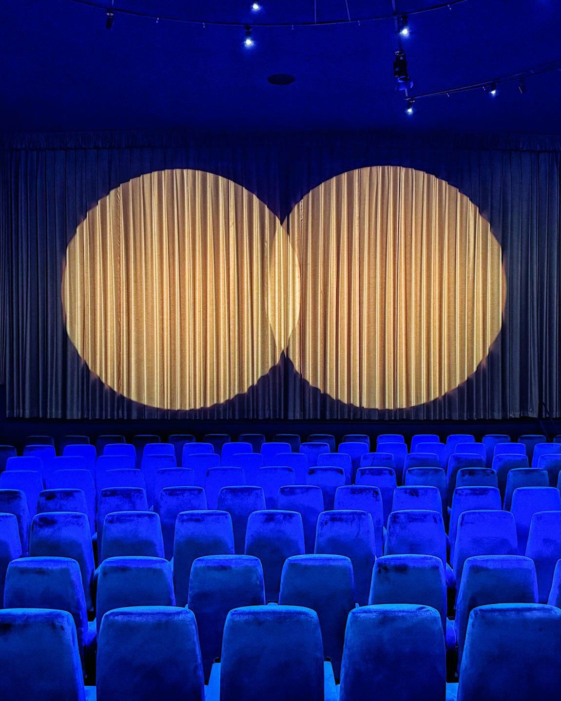 Interior of a dark cinema with cobalt blue velvet seats and two spotlights on a closed curtain covering the screen