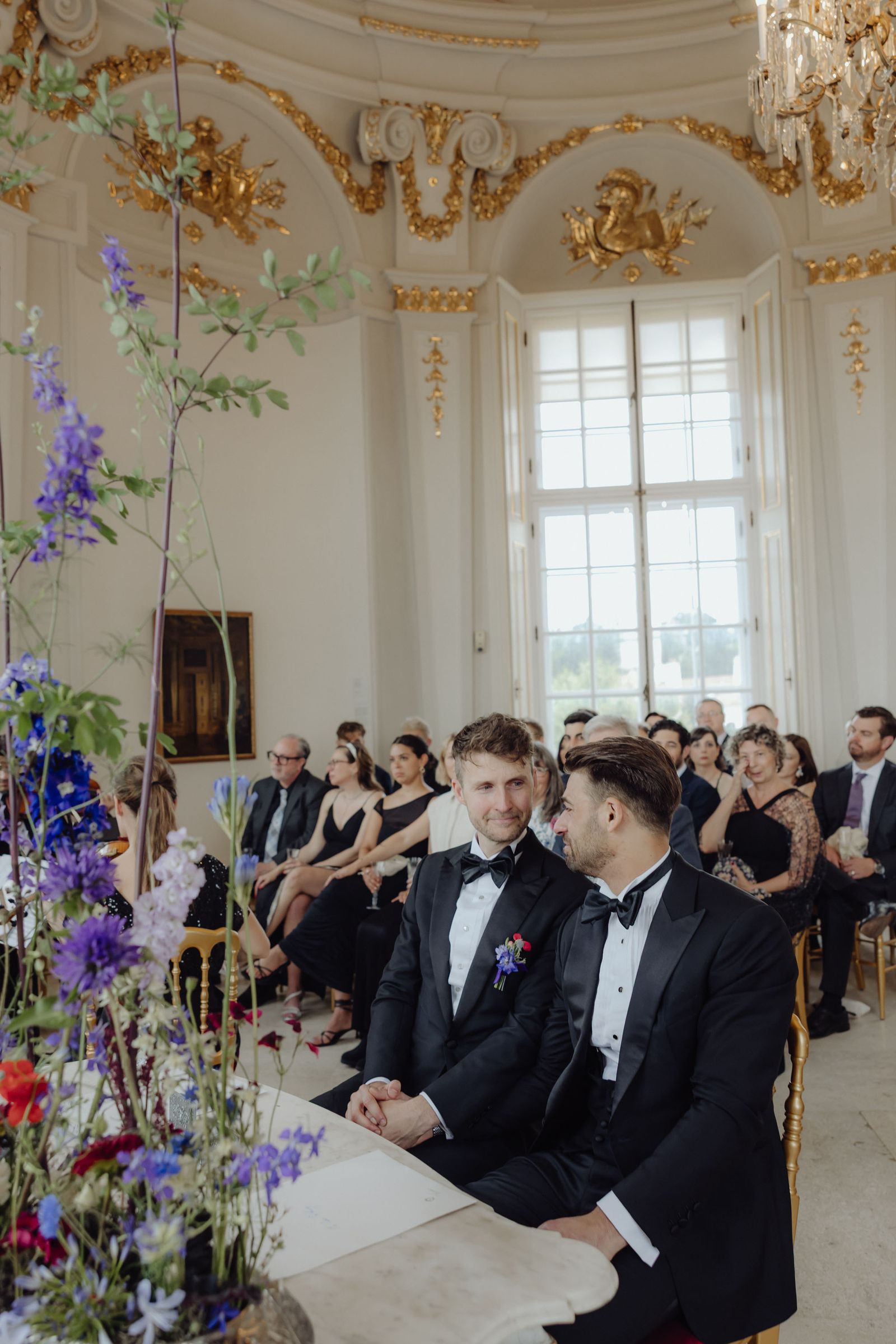 Two people sitting next to each other at the front of an ornate white room with gold accents and an audience behind them