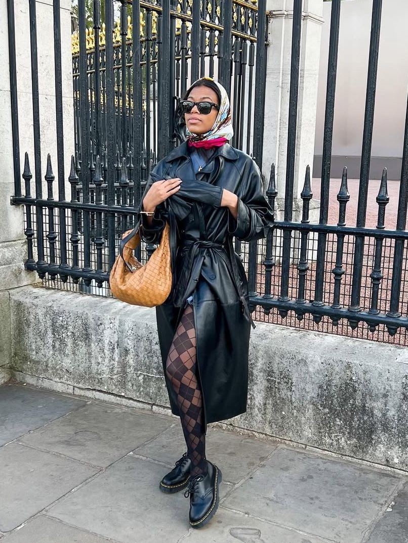 Person wearing black leather trench coat patterned tights a head scarf and brown leather bag while posing in front of an...