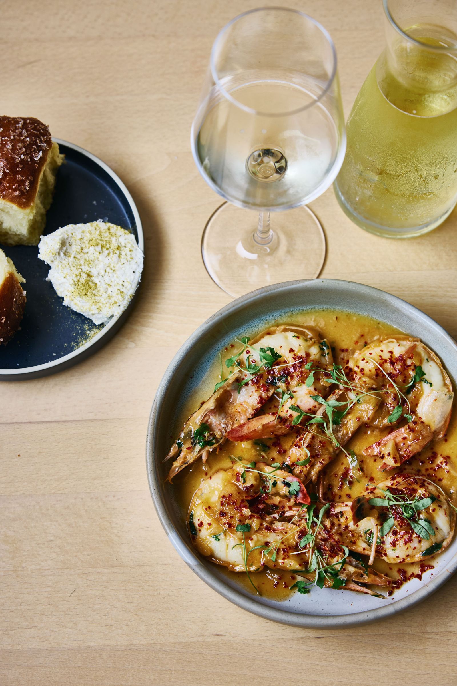 Photograph of a shrimp dish with a side of breach and a glass of white wine on a wooden surface
