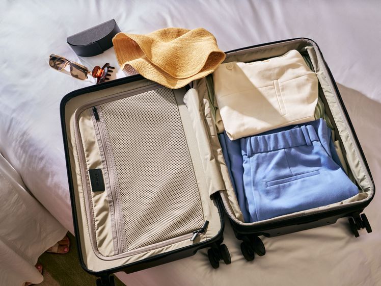Open suitcase with folded clothes inside and a straw hat and various accessories laying nearby